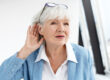 Senior woman in blue jacket cupping her ear, appearing to have trouble hearing.