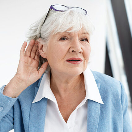 Senior woman in blue jacket cupping her ear, appearing to have trouble hearing.
