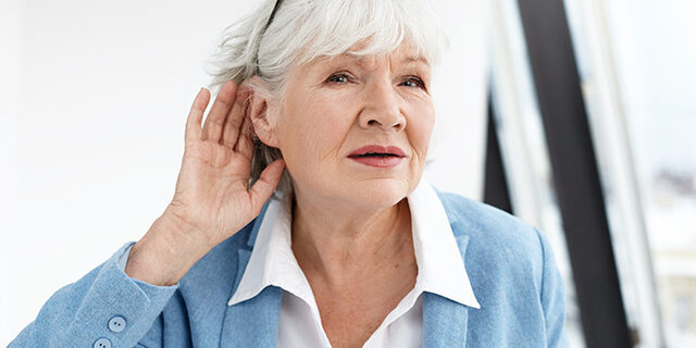 Senior woman in blue jacket cupping her ear, appearing to have trouble hearing.
