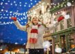 Woman in winter clothes smiles joyfully under festive lights and falling snow in a city street.